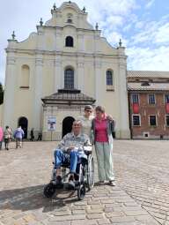 Na zdjęciu dwie mieszkanki stojące oraz mieszkaniec na wózku przed Bazyliką Krzyża Świętego w Mogile.
