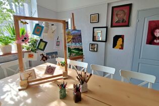 Na zdjęciu widać wnętrze pracowni plastycznej. Na środku stoi stół, na stole znajduje się drewniana rama na której rozwieszone są ręcznie wykonane kartki. W tle na ścianach widać obrazy, portrety.