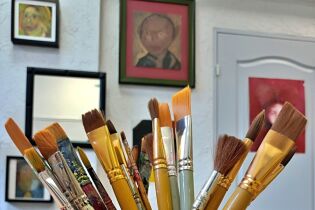 fotografia przedstawia na pierwszym planie pędzle malarskie stojące w naczyniu. W tle na ścianie widać obrazy. 