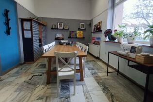 Fotografia przedstawia wnętrze pracowni rękodzieła. na środku stoi podłużny stół, wokół niego stoją krzesła. Na ścianach prezentowane są kolorowe prace wykonane przez uczestników warsztatów. 