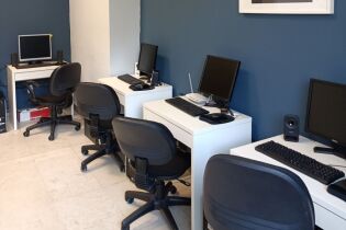 fotografia ukazuje wnętrze pracowni komputerowej. Widzimy biurka ustawione przy ścianach a na nich monitory i drukarkę. Przy biurkach stoją fotele biurowe.