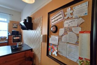 Fotografia przedstawia pomieszczenie w którym znajdują sie meble kuchenne i sprzęt AGD. Na pierwszym planie na ścianie wisi duża tablica z różnymi przepisami i notatkami