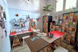 fotografia przedstawia pomieszczenie w którym powstają witraże. Przy ścianach znajdują się wielobarwne meble a na środku stół. Wszędzie poukładane i porozwieszane są różnej wielkości witrażyki. 