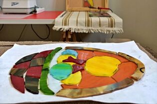 fotografia przedstawia kolorowe szkła ułożone w kształt koszyka wielkanocnego z jajkami i kurczątkiem w środku. Szkła leżą na stole i są przygotowane do zespolenia przy użyciu cyny. 