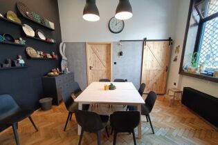 Fotografia przedstawia pomieszczenie z dużym stołem na środku. Wokół stołu znajdują się krzesła. Na ścianach powieszone są półki a na nich wyeksponowane różne wyroby ceramiczne.