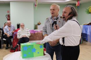 Dwaj stojący mężczyźni z mikrofonami. Obok na stoliku leży pudełko. W tle siedzący mężczyźni.