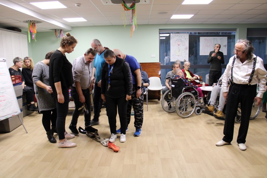 Grupa kobiet i mężczyzn w pomieszczeniu. Na pierwszym planie trzy kobiety i trzech mężczyzn podąża wzrokiem za ułożonymi w prostej linii butami. 