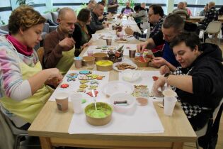 Grupa kobiet i mężczyzn zgromadzonych przy stołach dekorujących pierniczki. 