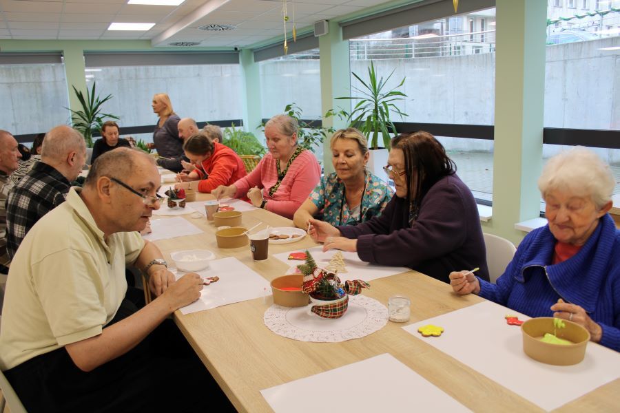 Grupa kobiet i mężczyzn siedzących przy stołach nakłada lukier na ciasteczka. 