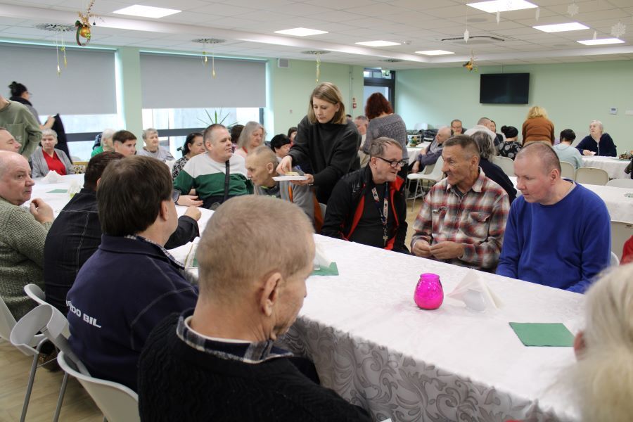 Grupa siedzących kobiet i mężczyzn przy nakrytych odświętnie stołach, w tle dekoracje bożonarodzeniowe. 