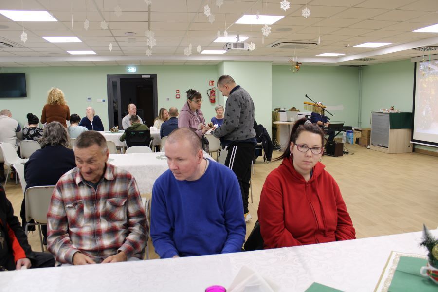 Grupa siedzących kobiet i mężczyzn przy nakrytych odświętnie stołach. Pośrodku stoi para, która łamie się opłatkiem.