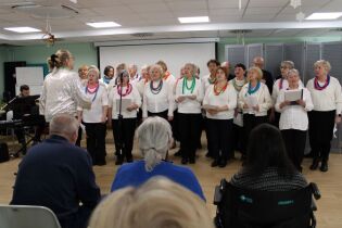 Grupa kobiet i mężczyzn stojących w dwóch rzędach. Przed nimi dwie kobiety i mężczyzna siedzący na widowni.