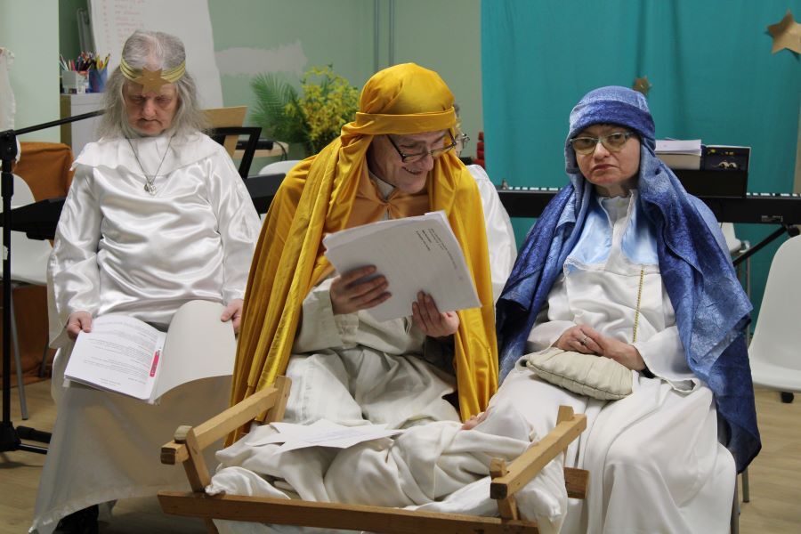 Grupa osób w kostiumach teatralnych występuje przed publicznością na tle świątecznej dekoracji.