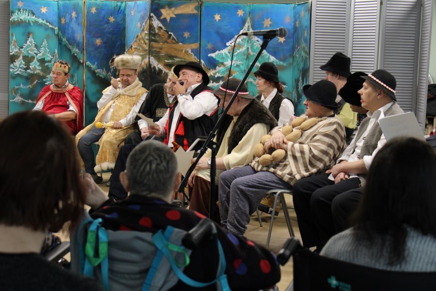 Grupa osób w pasterskich i góralskich strojach siedzi  na scenie, odgrywając scenę z jasełek.