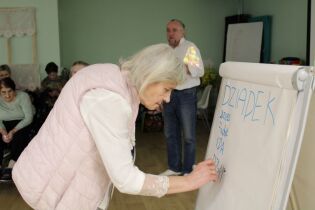 Kobieta pisze na tablicy, w tle grupa osób siedzi i obserwuje.