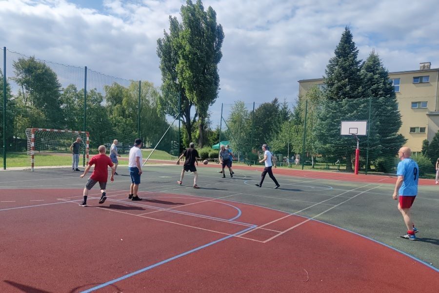 Grupa mężczyzn gra w piłkę nożną na boisku. W tle kosz do koszykówki, budynek i drzewa.