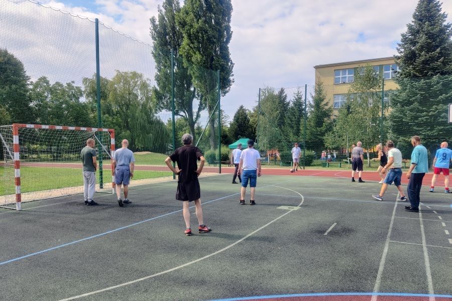 Grupa mężczyzn gra w piłkę nożną na boisku sportowym. W tle budynek oraz drzewa.