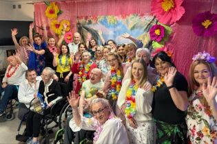 duża grupa osób machających rękoma. Większość z nich na szyi ma girlandy z kwiatów w stylu hawajskim