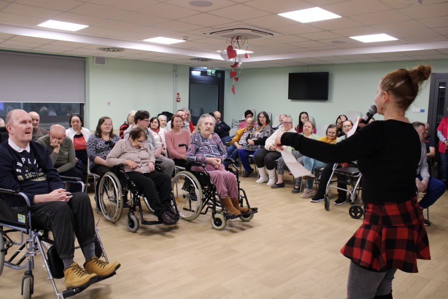 Kobieta z mikrofonem prowadzi spotkanie w pomieszczeniu. Przed nią siedzi publiczność, w tym osoby na wózkach inwalidzkich. 