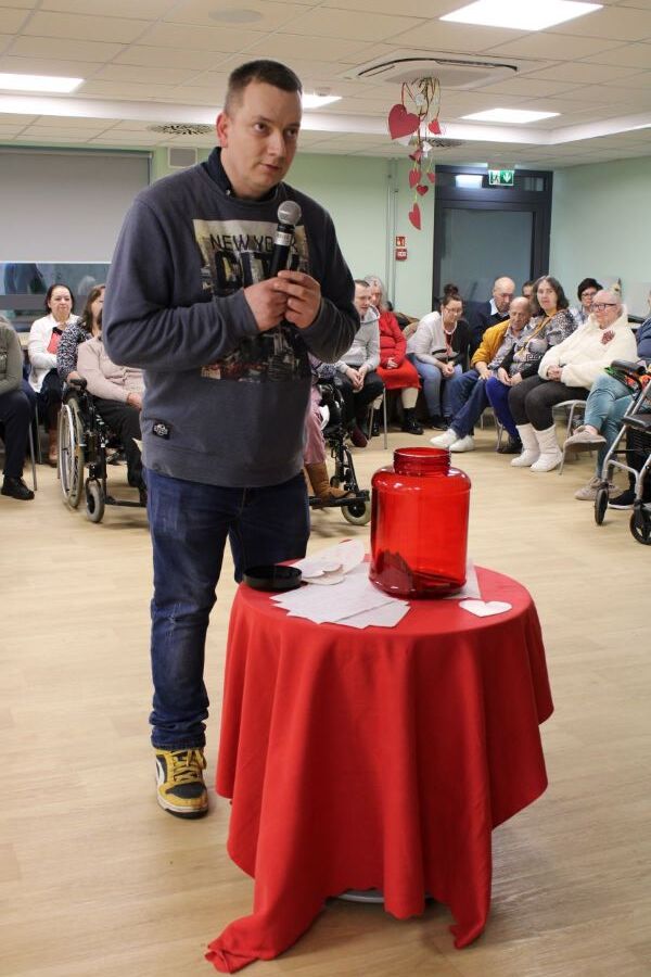 Mężczyzna z mikrofonem stoi przy stoliku przykrytym obrusem. Na stole znajduje się duży słoik oraz kartki w kształcie serc. W tle widownia.