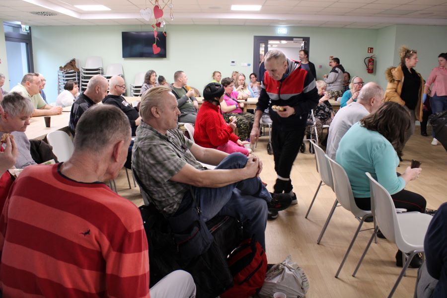 Grupa osób, w tym mężczyzna na wózku inwalidzkim, siedzą w rzędzie na krzesłach. W tle pozostali uczestnicy spotkania.