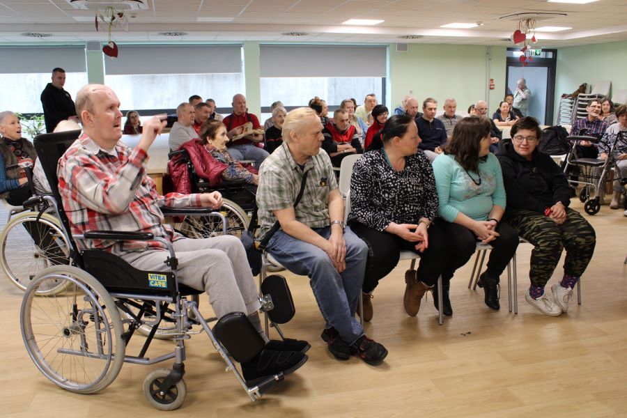 Grupa osób, w tym mężczyzna na wózku inwalidzkim, siedzą w rzędzie na krzesłach. W tle pozostali uczestnicy spotkania.