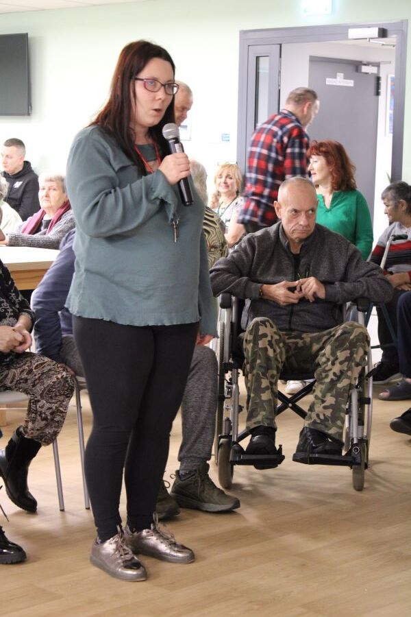 Kobieta w okularach stoi z mikrofonem w ręku. W tle widownia.