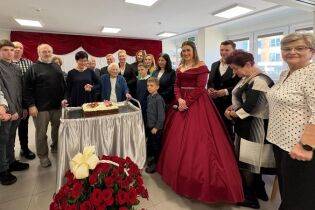 Grupa osób odświętnie ubranych stoi obok tortu. po środku stoi starsza kobieta, jubilatka