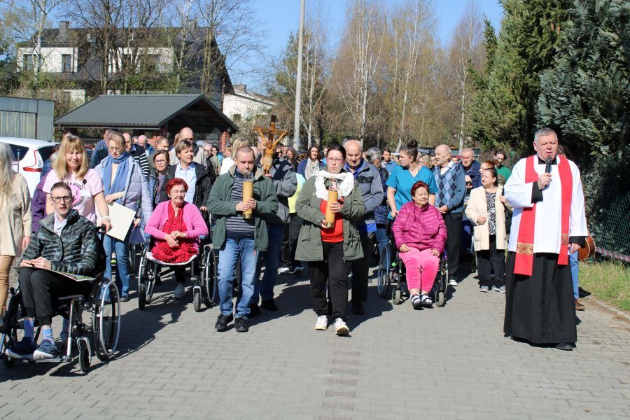 Grupa osób uczestniczy w nabożeństwie Drogi Krzyżowej na świeżym powietrzu. Na czele procesji idą osoby niosące świece oraz ksiądz z mikrofonem. W tle budynki i drzewa
