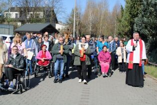 Grupa osób uczestniczy w nabożeństwie Drogi Krzyżowej na świeżym powietrzu. Na czele procesji idą osoby niosące świece oraz ksiądz z mikrofonem. W tle budynki i drzewa
