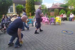 Grupa osób bierze udział w zabawie na świeżym powietrzu; uczestnicy rzucają woreczkami do celu przy dopingu widowni. 