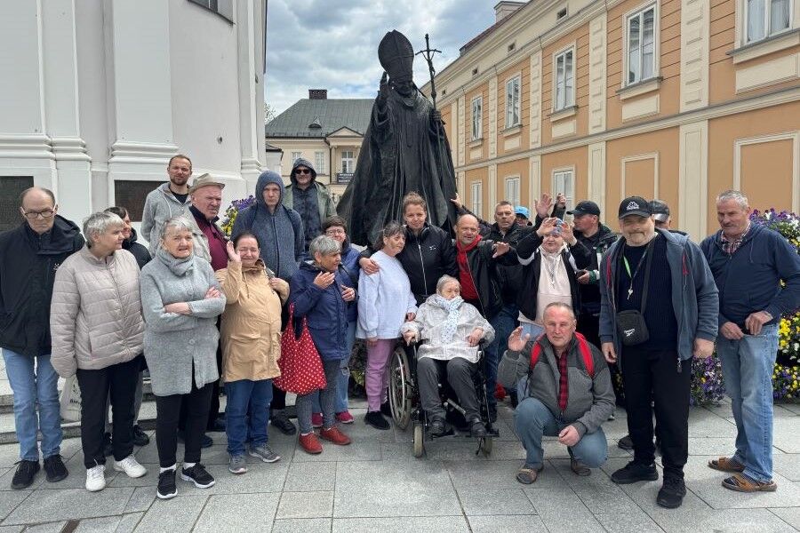Grupa osób pozuje przed pomnikiem, w tle zabytkowe budynki oraz kwietniki.