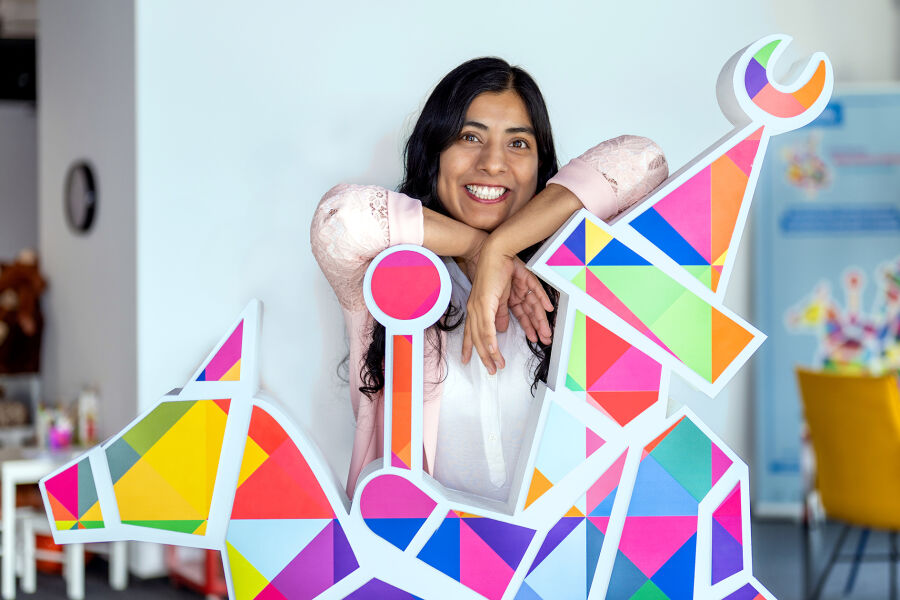 Długowłosa, ciemnowłosa dziewczyna o ciemnej karnacji pozuje oparta o dużą, kolorową figurę krakowskiego Lajkonika. Dziewczyna jest ubrana w różową bluzkę. Uśmiecha się szeroko. 