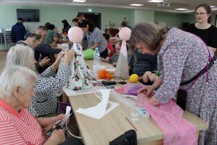 Grupa kobiet i mężczyzn intensywnie pracują przy wspólnym stole, dekorując anioły tkaninami i włóczką. Panuje twórcza, skupiona atmosfera.