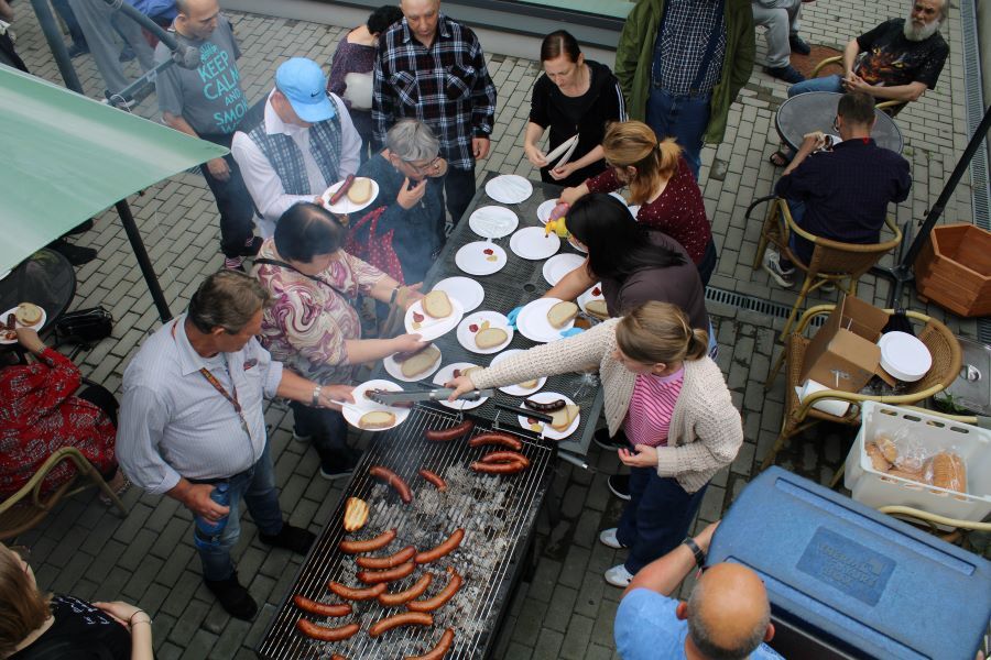 Grupa osób zebrana przy dużym grillu z kiełbaskami na świeżym powietrzu. Przygotowywany jest posiłek, rozkładane są talerze z chlebem, musztardą i keczupem. Część osób siedzi przy stolikach.