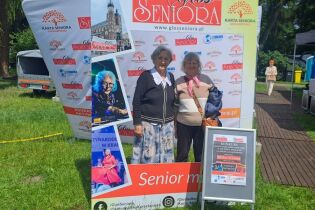 Zdjęcie prezentuje seniorki na tle stendu organizatora Stowarzyszenia Manko – Głos Seniora