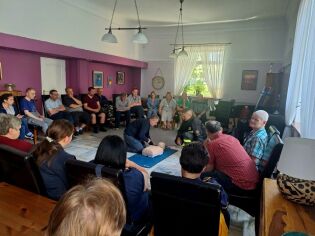 Uczestnicy siedzą w grupie i obserwują, jak jedna z osób ćwiczy odpowiednie zachowanie podczas reanimacji  na warsztatach z pierwszej pomocy