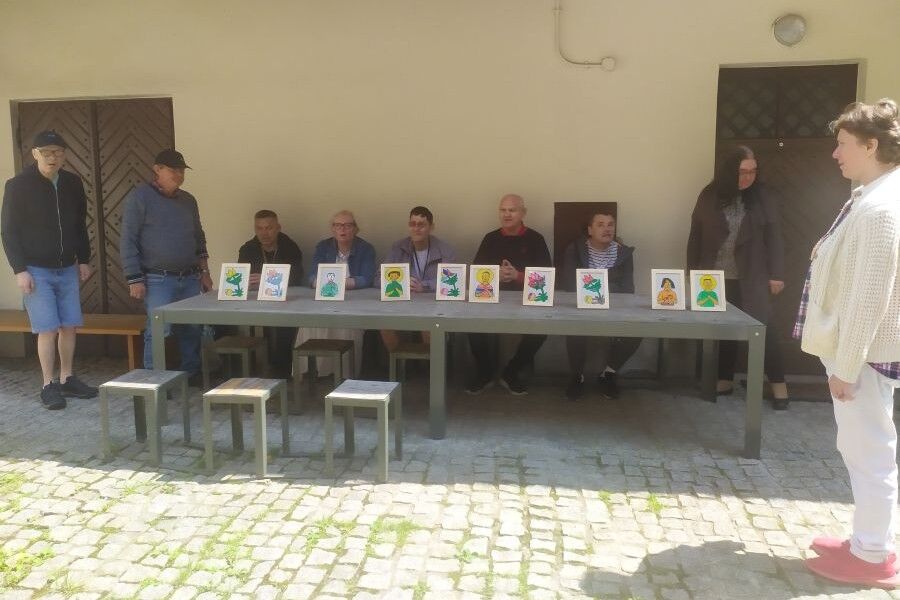 Trzy kobiety i sześciu mężczyzn zgromadzonych przy prostokątnym stole, na którym ustawiono kolorowe obrazki w ramkach. Grupa prezentuje swoje prace, na tle dziedzińca z brukowaną nawierzchnią.
