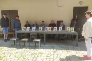 Trzy kobiety i sześciu mężczyzn zgromadzonych przy prostokątnym stole, na którym ustawiono kolorowe obrazki w ramkach. Grupa prezentuje swoje prace, na tle dziedzińca z brukowaną nawierzchnią.