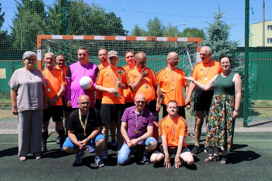 Zwycięska drużyna piłkarska stojąca przed bramką, pozująca do wspólnego zdjęcia. Wszyscy mają na szyjach medale, a jeden z zawodników trzyma trofeum. Po bokach stoją dwie kobiety.