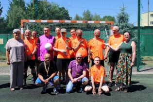 Zwycięska drużyna piłkarska stojąca przed bramką, pozująca do wspólnego zdjęcia. Wszyscy mają na szyjach medale, a jeden z zawodników trzyma trofeum. Po bokach stoją dwie kobiety.