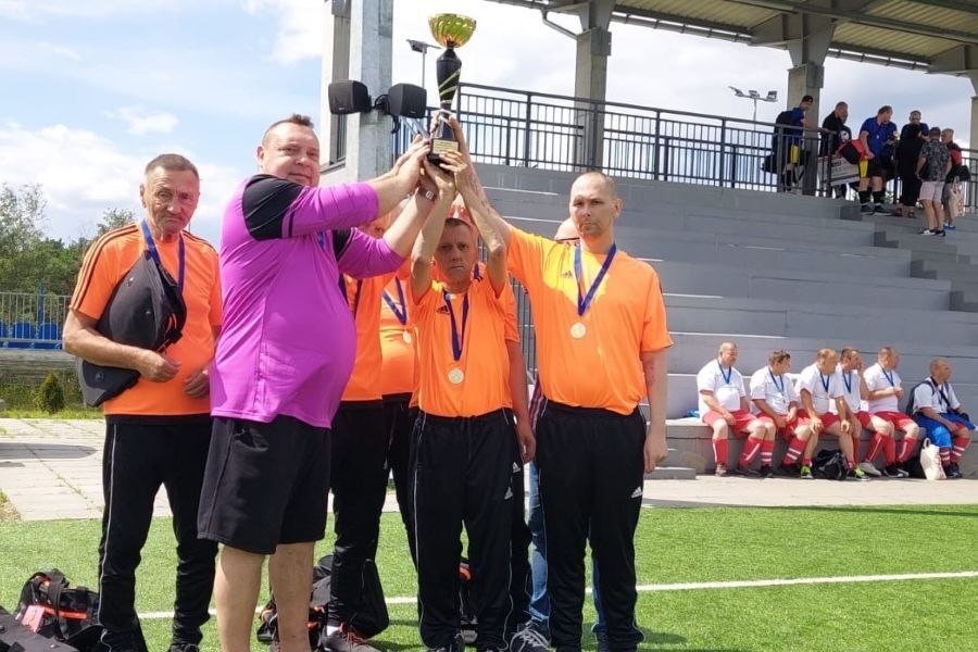 Zwycięska drużyna piłkarska pozuje do zdjęcia po zakończonym turnieju. Mężczyźni trzymają trofea i medale, a w tle ustawione są flagi z logo turnieju. 