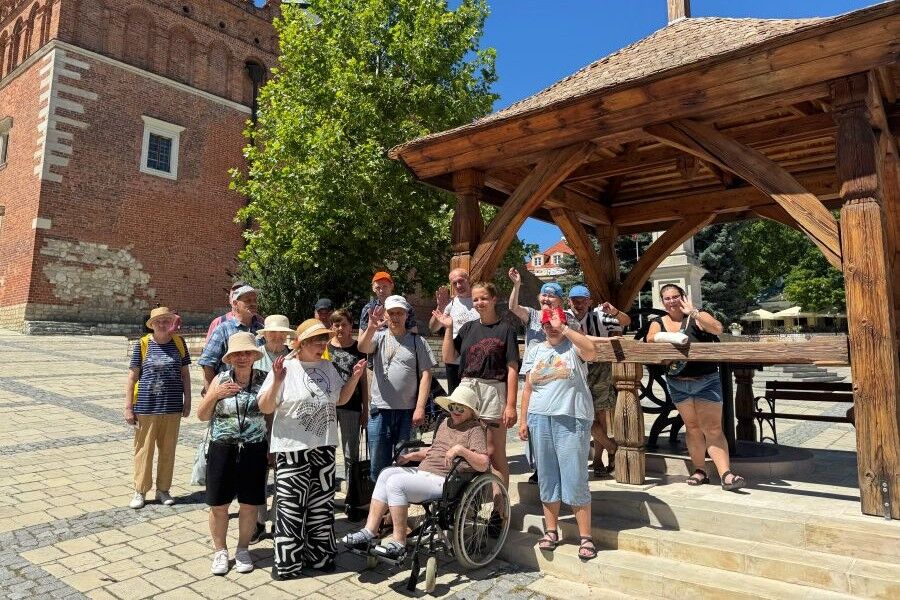Grupa kobiet i mężczyzn stoi i pozuje do pamiątkowej fotografii przy drewnianej studni na tle ceglanego kościoła. Jest słoneczny dzień, uczestnicy wycieczki machają i uśmiechają się do zdjęcia.