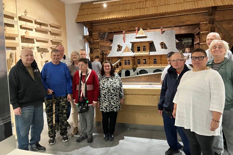 Grupa zwiedzających pozuje wewnątrz muzeum przy ekspozycji przedstawiającej model tradycyjnego góralskiego domu z drewna. Na bocznej ścianie wiszą drewniane detale architektoniczne.