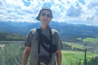 Mężczyzna w ciemnych okularach przeciwsłonecznych, czapce z daszkiem przekrzywionej do tyłu i sportowej odzieży stoi na tle panoramy gór. 