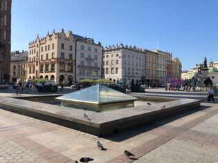 Zdjęcie przedstawia fontannę na Rynku Głównym w Krakowie. Oszklona fontanna w kształcie piramidy, w tle kamienice oraz pomnik Adama Mickiewicza.
