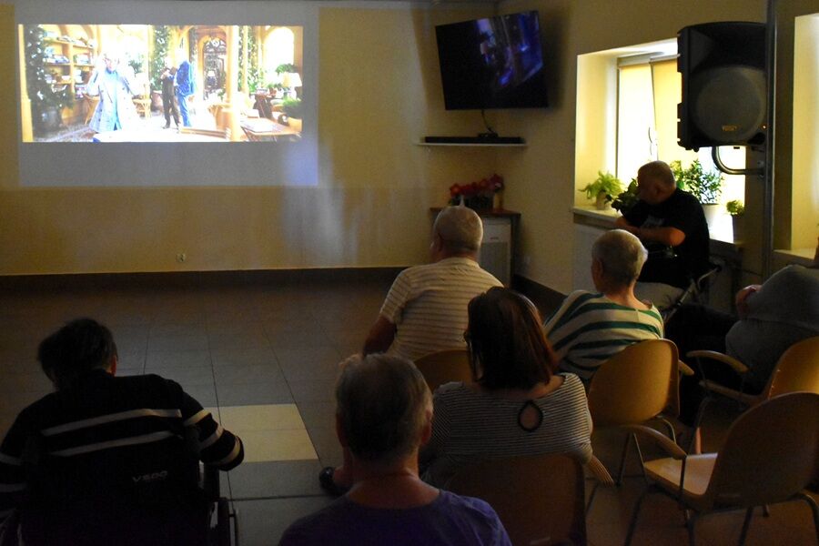 Grupa osób siedzi na krzesłach w sali i ogląda film wyświetlany na dużym ekranie na ścianie. Widać ciemne pomieszczenie, ekran z kolorową sceną filmową oraz ludzi oglądających projekcję.