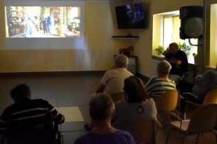 Grupa osób siedzi na krzesłach w sali i ogląda film wyświetlany na dużym ekranie na ścianie. Widać ciemne pomieszczenie, ekran z kolorową sceną filmową oraz ludzi oglądających projekcję.