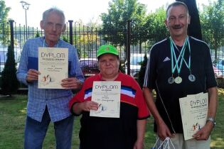 Trzy osoby stoją na świeżym powietrzu, trzymając dyplomy za udział i miejsca w zawodach sportowych. Dwóch mężczyzn i jedna kobieta uśmiechają się do zdjęcia.