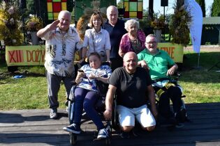 Grupa osób, w tym osoby starsze i osoby z niepełnosprawnościami, pozuje razem na tle sceny ozdobionej kwiatami i kolorowymi elementami. Część osób siedzi na wózkach inwalidzkich, inni stoją za nimi.